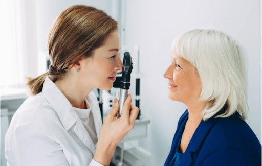 Young female optometrist examining a mature female patient with white hair with an opthalmascope to check for retinal concerns.