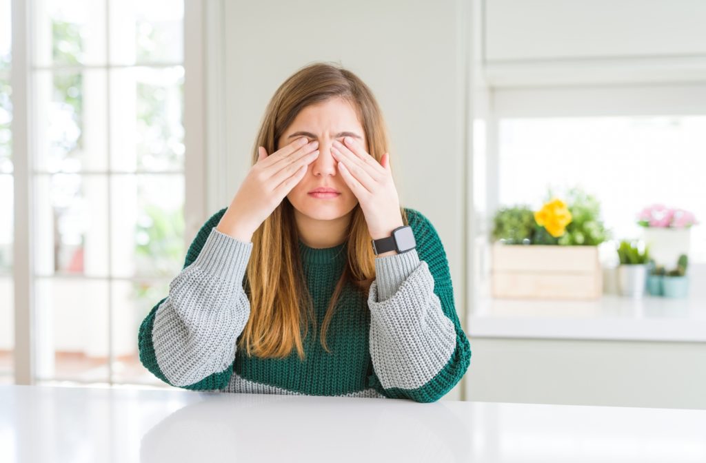 A woman leaning on her white kitchen counter holding her eye's as she is experiencing a burning sensation due to dryness