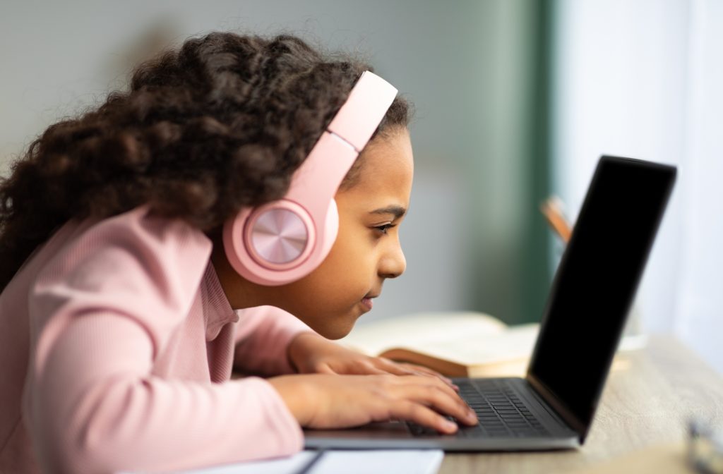 A young girl squinting with pink headphones on, looking at her laptop extremely closely as she is struggling to read the text on the screen