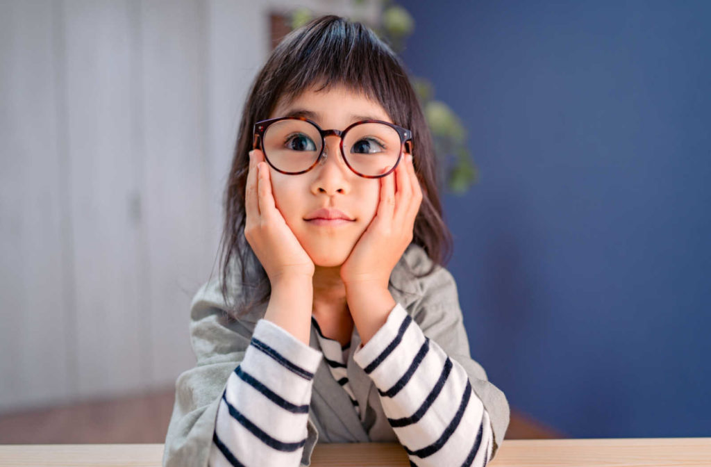 A young girl with a lazy eye wearing glasses with her hands on her cheeks.
