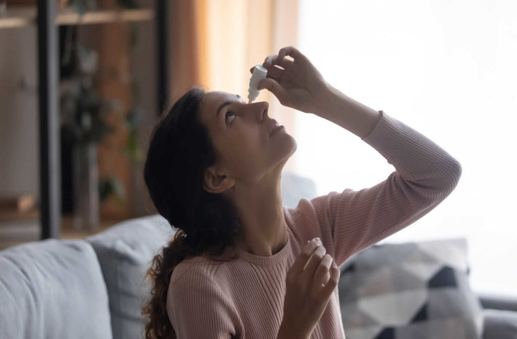 A woman sitting on her couch applying eye drops to her eye to help ensure she doesn't rub her eyes and cause damage to the cornea.