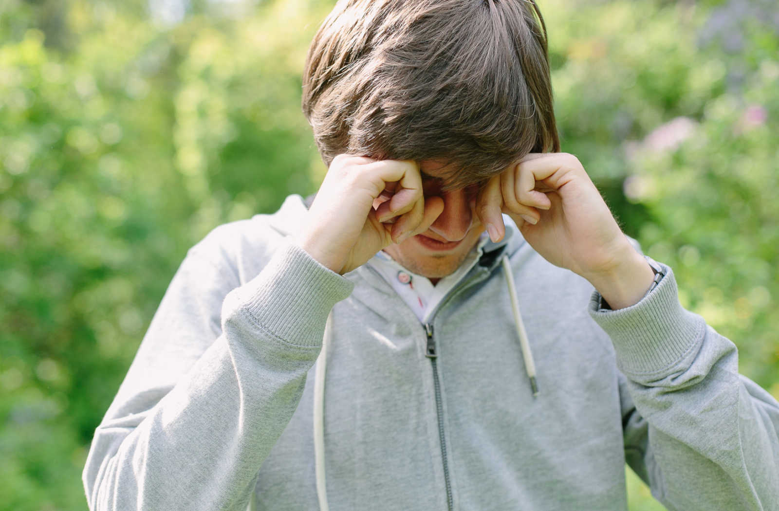 A young male standing outside rubbing his eyes.