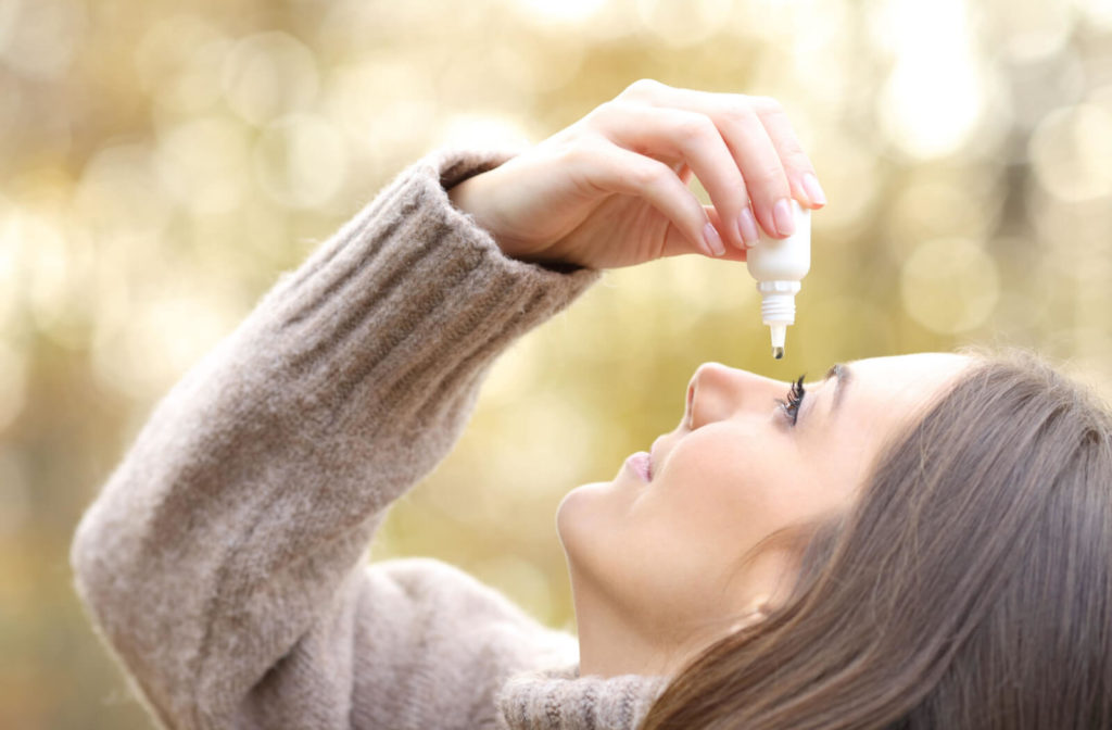 A woman in a brown sweatshirt is holding a small white bottle in her right hand  and dropping  an artificial  tear on her left eye