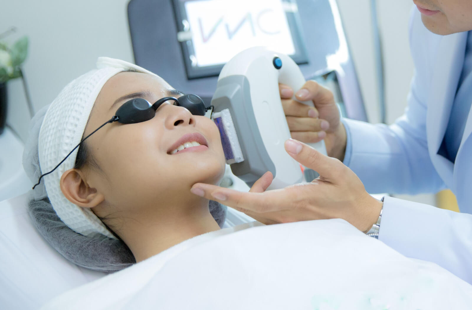 A smiling, reclined patient wearing goggles receiving IPL treatments on her face from her optometrist