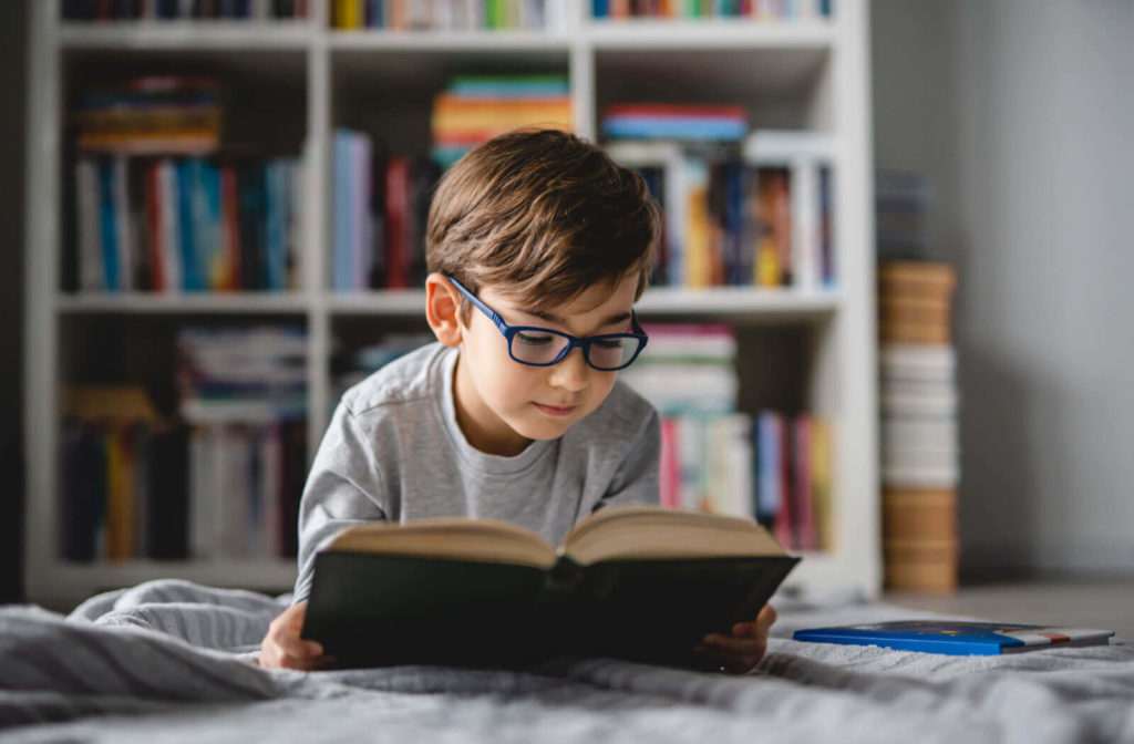 A boy with prescription eyeglasses reading a book comfortably.