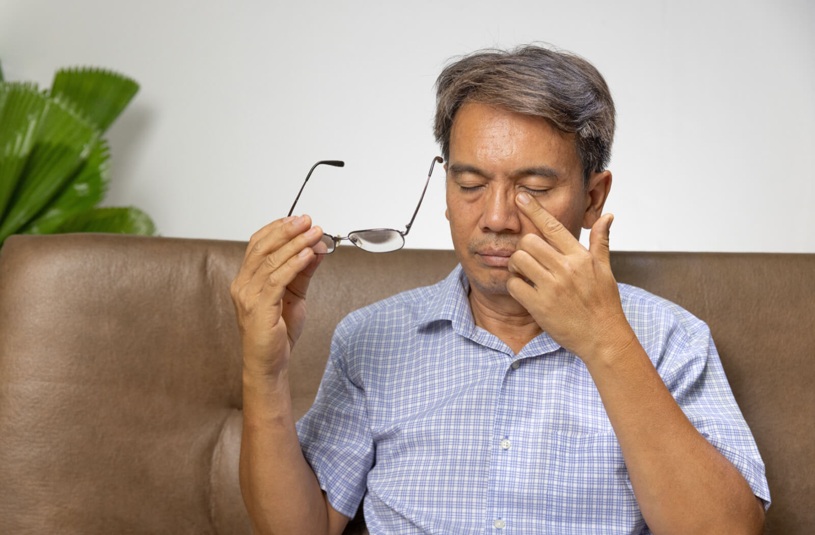 A senior man sitting on a couch and holding his glasses in his right hand as he rubs his left eye with his left hand.