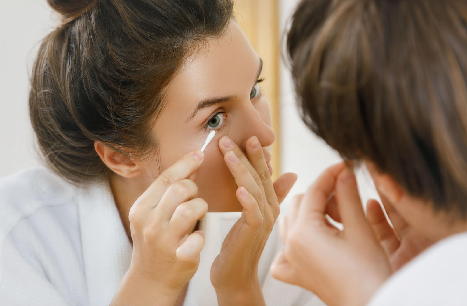 A youthful lady is gently swabbing her eyelids with a cotton swab while standing before a mirror.
