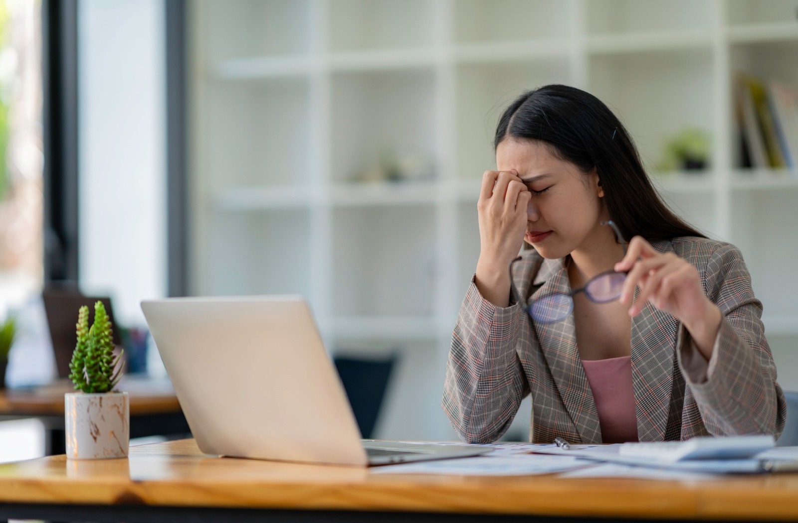 A professional-looking woman working at a laptop winces in pain due to digital eye strain