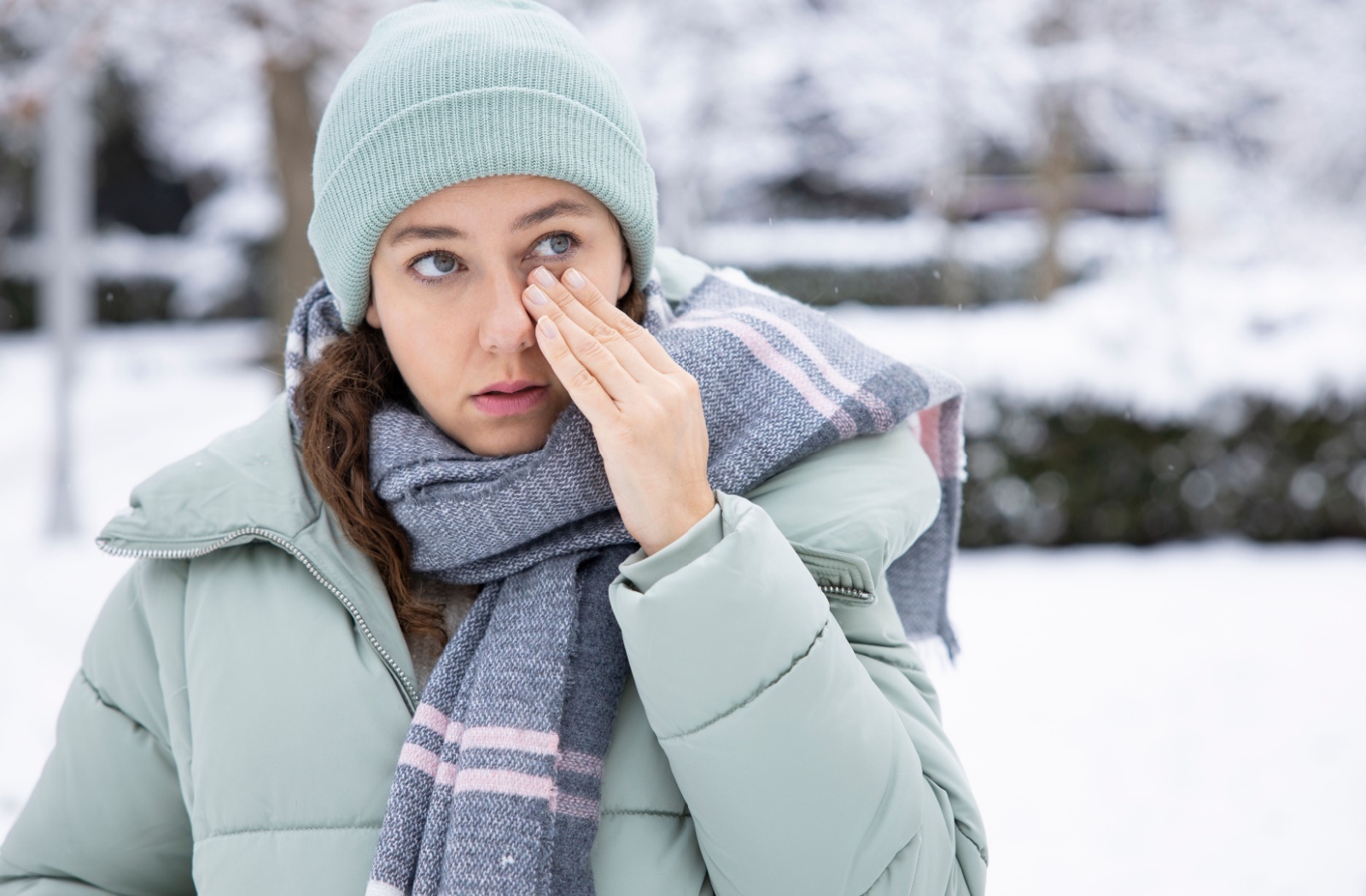 A person in winter clothing stands outside during a winter day. They're rubbing one eye, suggesting that eye is dry or irritated.