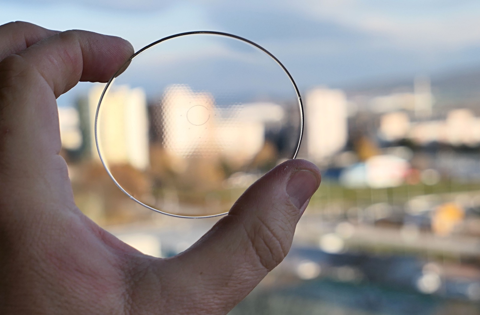 A close-up of a hand holding a MiyoSmart lens up to a cityscape.
