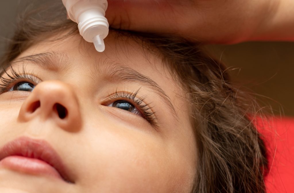 A close-up image of a child lying down while their parent applies eye drops to their left eye.