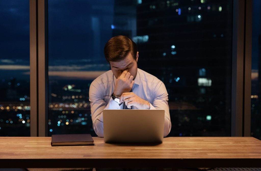 A professional-looking young man rubs at both of his eyes after a long day working on his laptop.