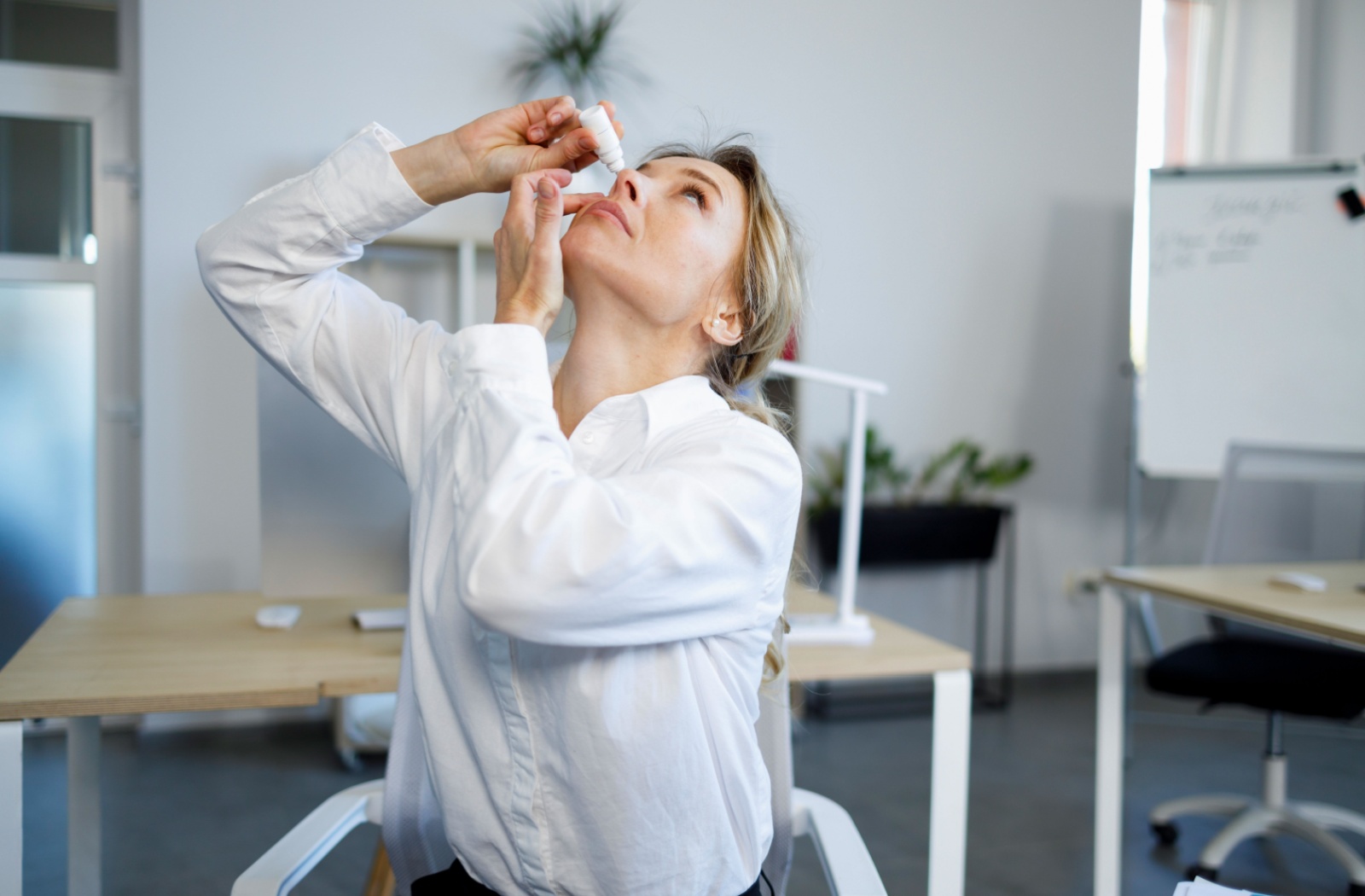 A young woman in an office space applies eye drops to her right eye.
