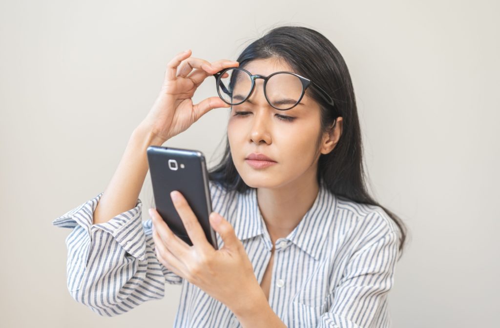 An adult lifting their glasses while trying to read their phone, struggling due to poor vision.