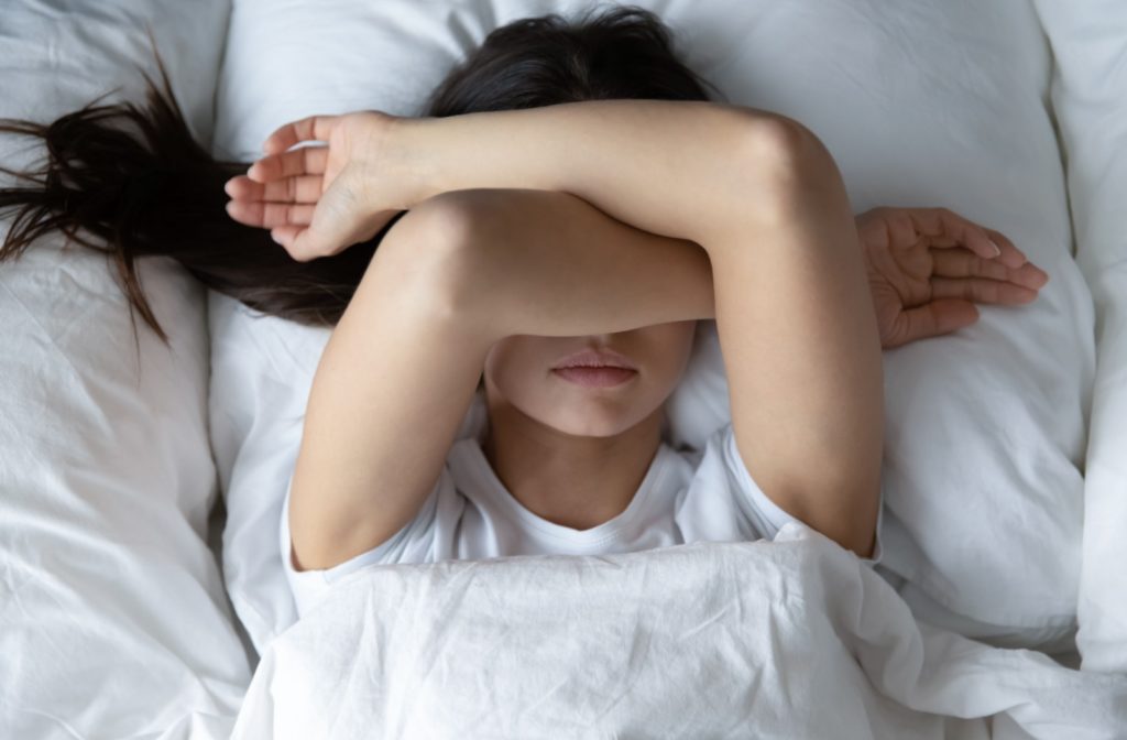 A young adult lying in bed with their forearms crossed over their eyes due to waking up with dry eyes.