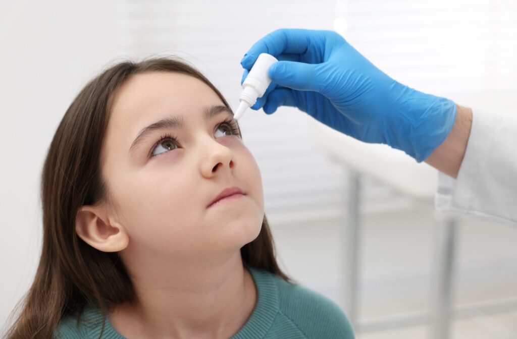 A child looks up as a gloved hand gently applies eye drops during an eye exam.