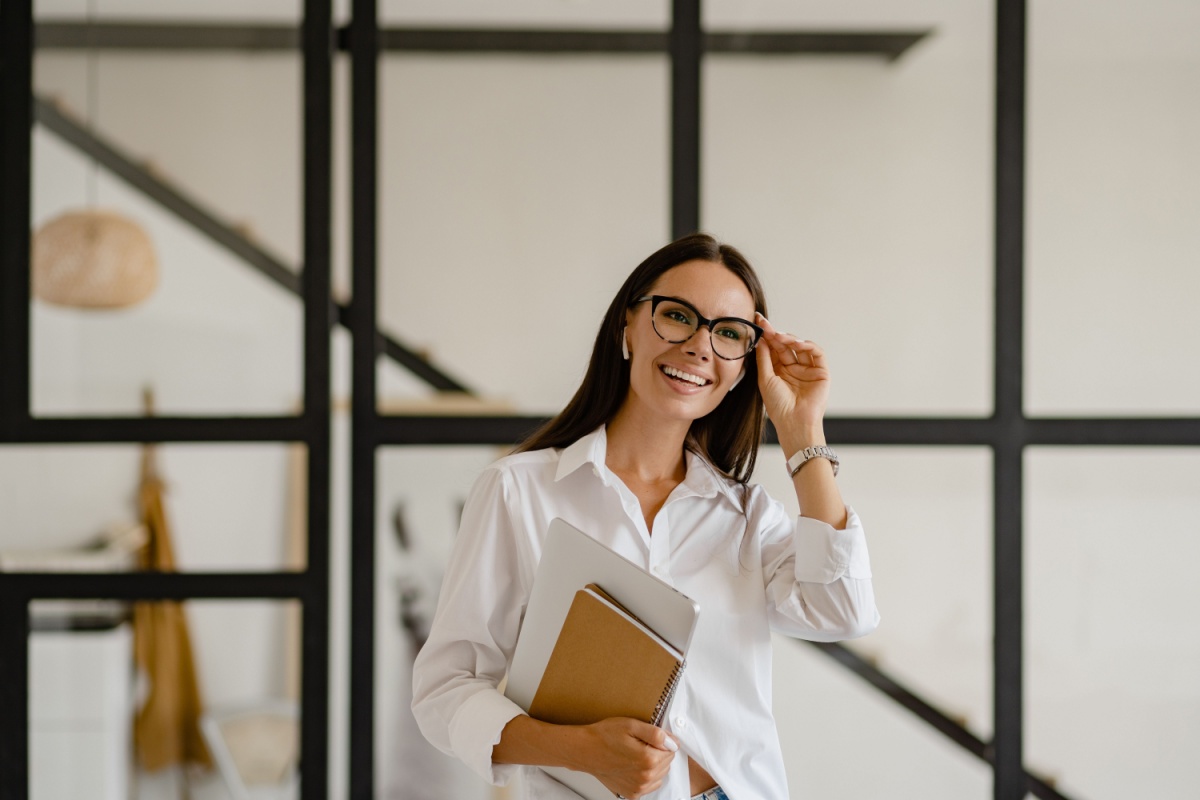 Confident businesswoman wearing prescription eyeglasses holding notebooks in modern office setting.