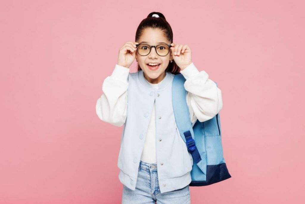 A cheerful young girl with a high ponytail wears a light blue varsity jacket and carries a blue backpack. She is smiling excitedly while holding up a pair of stylish, round-framed glasses to her face, posing against a solid pink background, symbolizing the joy of clear vision for back-to-school.