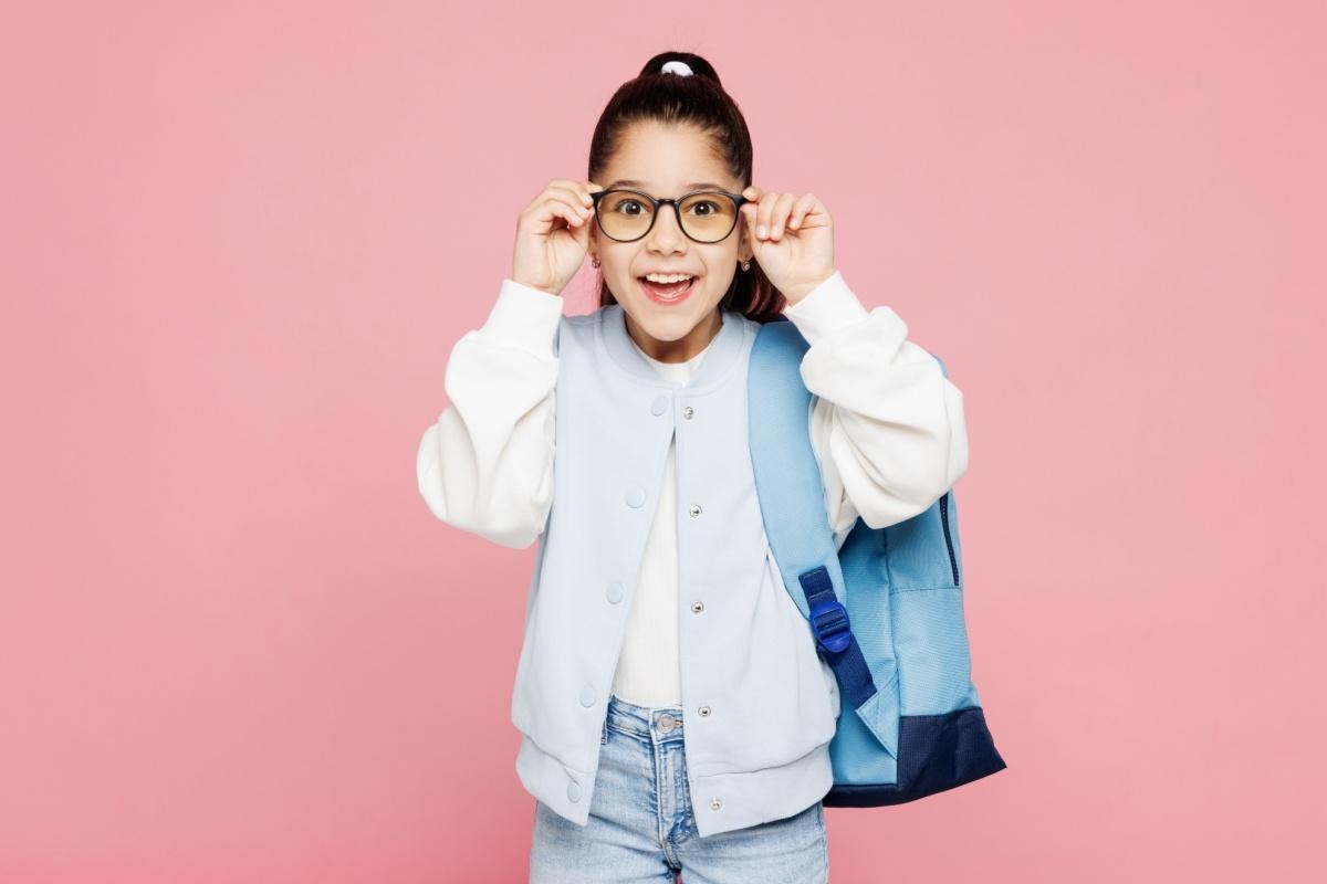 A cheerful young girl with a high ponytail wears a light blue varsity jacket and carries a blue backpack. She is smiling excitedly while holding up a pair of stylish, round-framed glasses to her face, posing against a solid pink background, symbolizing the joy of clear vision for back-to-school.
