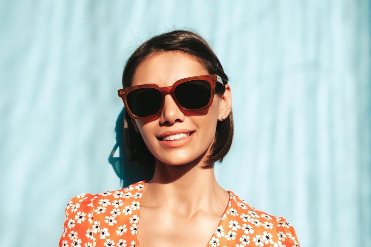 A smiling young woman with short dark hair wearing retro-style brown sunglasses against a bright blue backdrop, showcasing fashionable and effective eye protection.