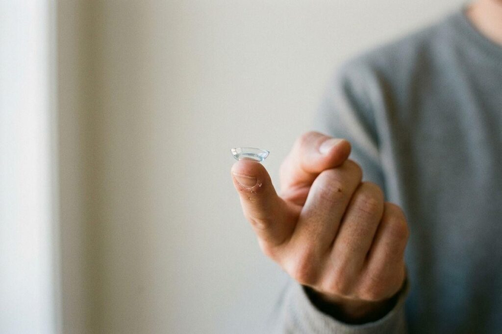 A contact lens balanced on a fingertip, held up by a person in a grey top against a plain light background.