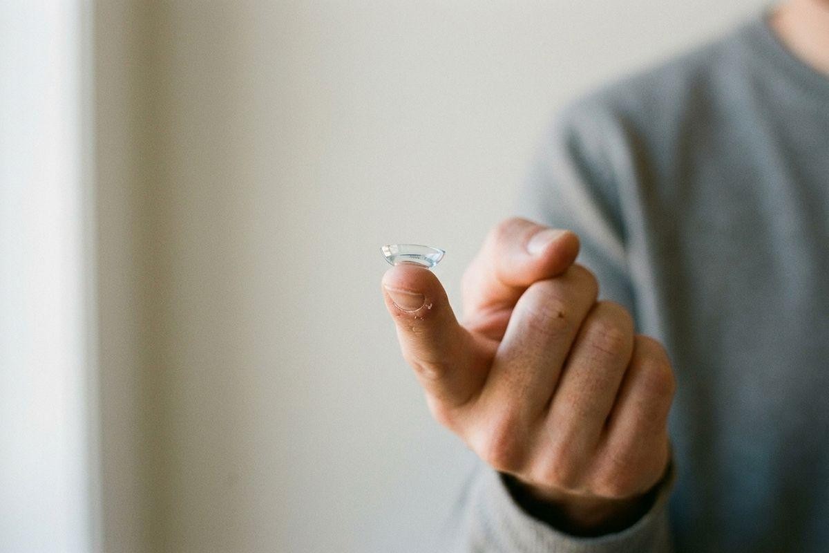 A contact lens balanced on a fingertip, held up by a person in a grey top against a plain light background.